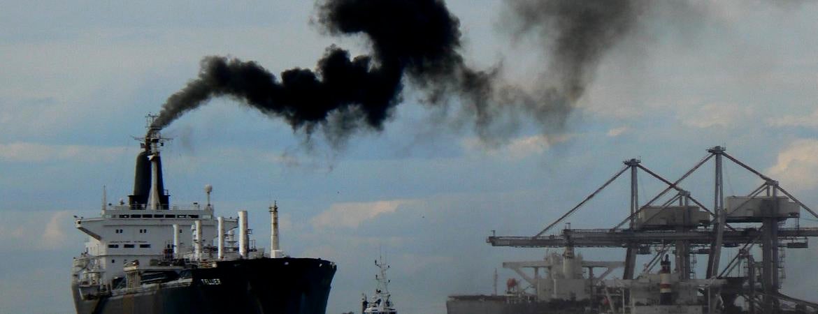 A cargo ship maneuvers out of port with black smokestack emissions