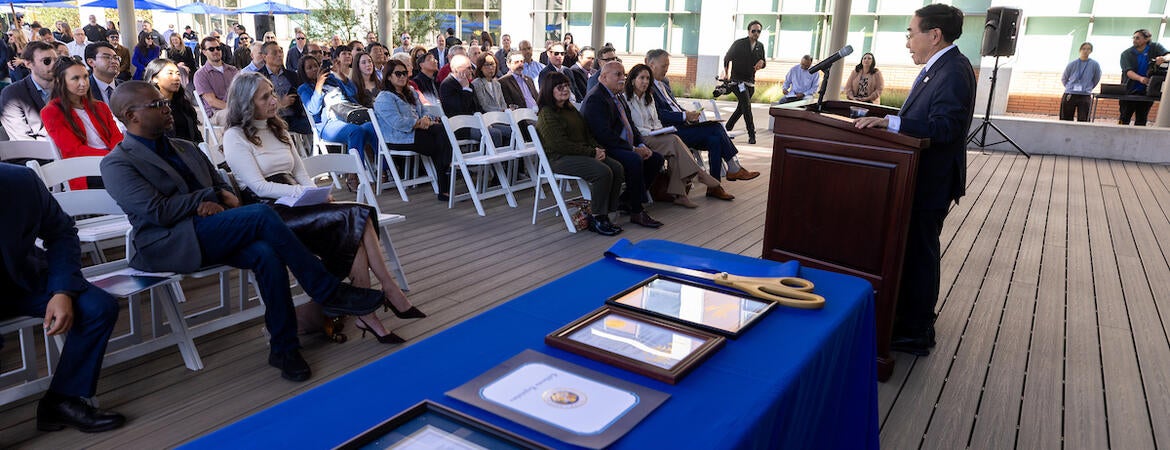 Man speaks at podium to outdoor audience; framed documents on blue table.