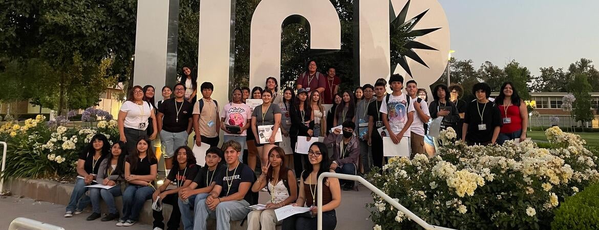 Group of young people by large UCR sign, belltower in background.