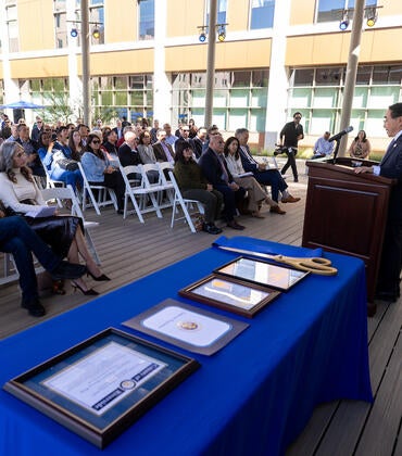Man speaks at podium to outdoor audience; framed documents on blue table.