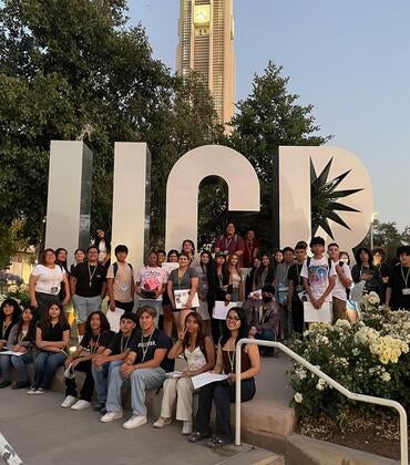 Group of young people by large UCR sign, belltower in background.