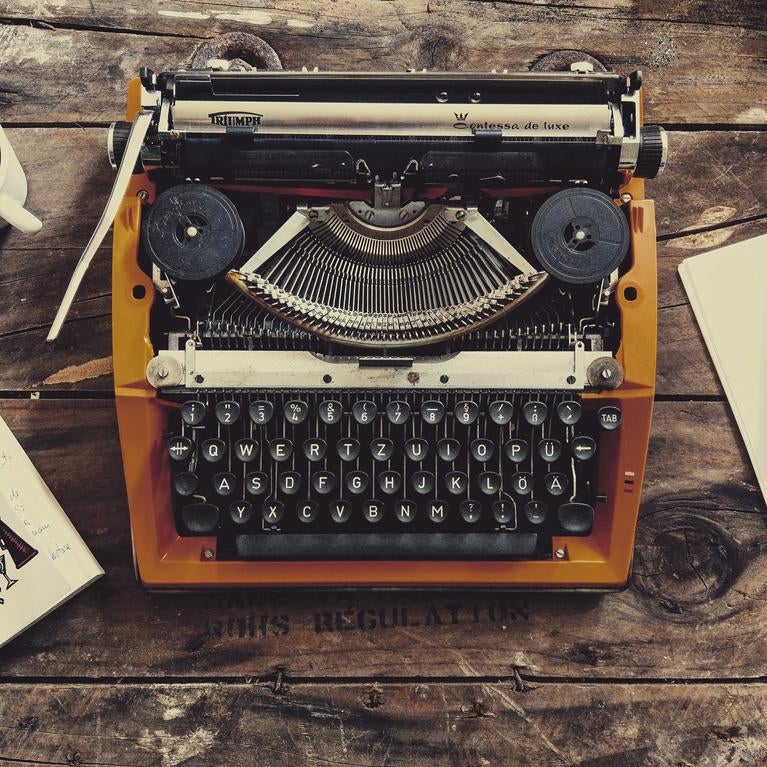 Typewriter on wooden desk with mug of coffee, book, paper, and phone.
