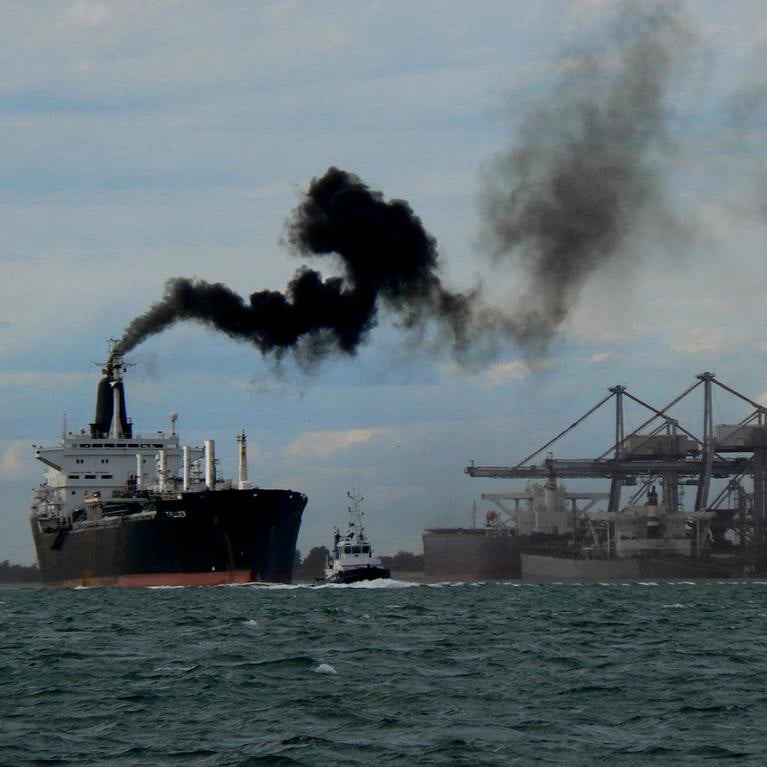 A cargo ship maneuvers out of port with black smokestack emissions