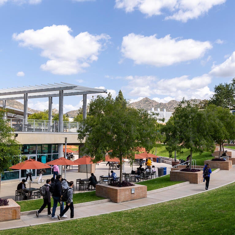 UCR campus: modern building, outdoor seating, green lawn, students, distant hills.