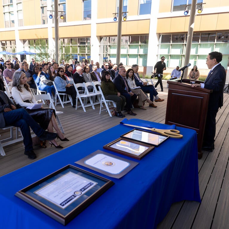 Man speaks at podium to outdoor audience; framed documents on blue table.