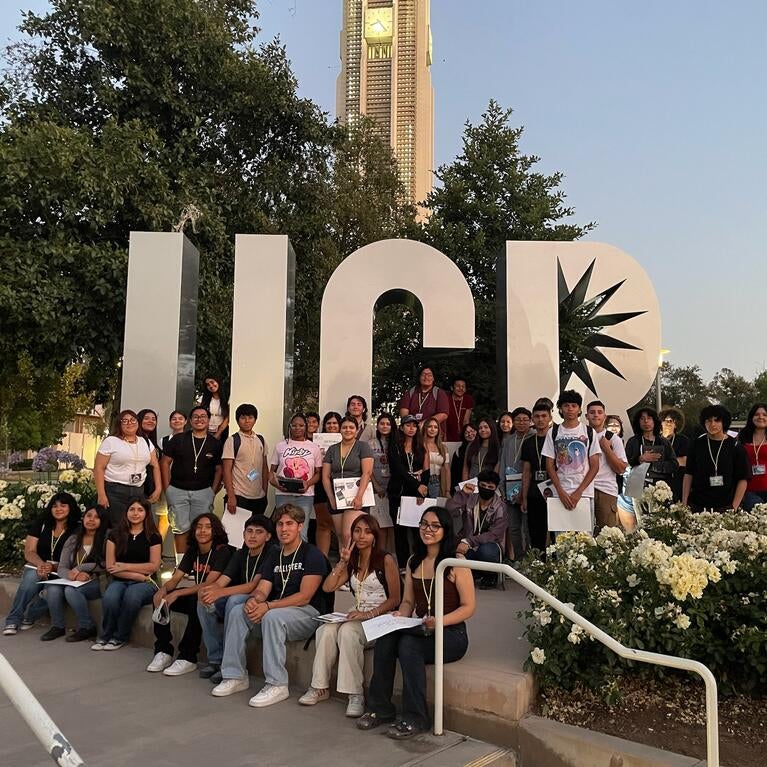 Group of young people by large UCR sign, belltower in background.