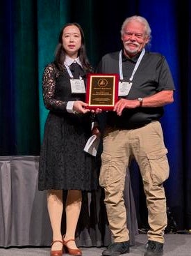 Yang Claire Yang and David Swanson stand next to each other, displaying the Mindel C. Sheps Award plaque that Swanson received.